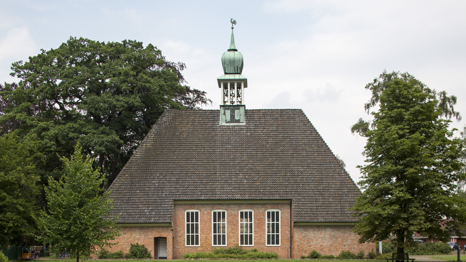 Eine rote Backsteinkirche mit einem hohen, spitzen Dach und einer kleinen Kupferkuppel. Vor der Kirche befinden sich grüne Wiesen und Bäume. Die Kirche hat große Fenster mit weißen Rahmen, die das Licht hereinlassen.