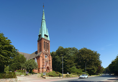 Eine rote Backsteinkirche mit einem hohen, spitzen Turm und einer Kupferhaube steht umgeben von Bäumen. Der Himmel ist blau und klar. Im Vordergrund verläuft eine Straße, auf der ein weißes Auto fährt.