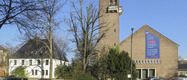 Ein Gebäude mit einem hohen Turm und einer großen Wandtafel, die einen Text über Liebe und Menschlichkeit zeigt. Die Wand ist mit einem blauen und roten Hintergrund gestaltet. Neben dem Gebäude steht ein weiteres, weißes Haus und Bäume sind im Vordergrund sichtbar.
