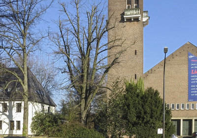 Ein Gebäude mit einem hohen Turm und einer großen Wandtafel, die einen Text über Liebe und Menschlichkeit zeigt. Die Wand ist mit einem blauen und roten Hintergrund gestaltet. Neben dem Gebäude steht ein weiteres, weißes Haus und Bäume sind im Vordergrund sichtbar.