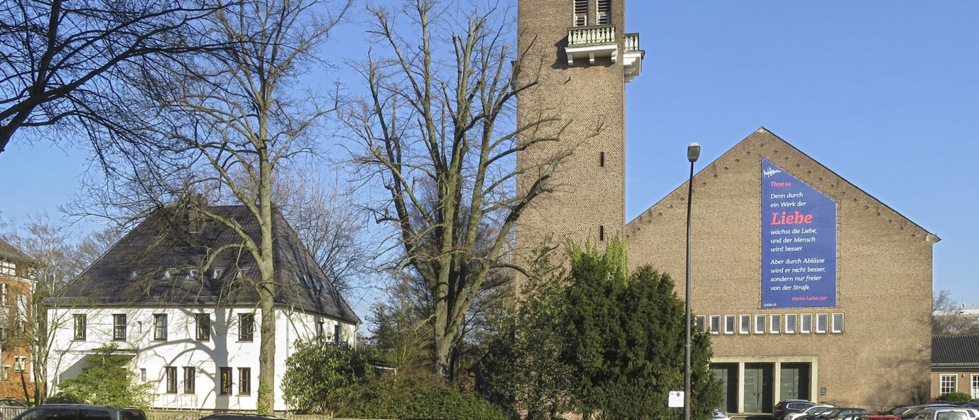 Ein Gebäude mit einem hohen Turm und einer großen Wandtafel, die einen Text über Liebe und Menschlichkeit zeigt. Die Wand ist mit einem blauen und roten Hintergrund gestaltet. Neben dem Gebäude steht ein weiteres, weißes Haus und Bäume sind im Vordergrund sichtbar.