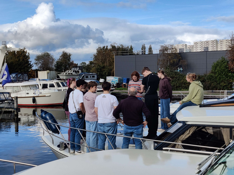 Boots-Einweisung Eine Gruppe von zehn Personen steht auf einem Boot im Hafen. Einige schauen auf ein Handy, während andere miteinander sprechen. Im Hintergrund sind weitere Boote und Bäume zu sehen, unter einem teilweise bewölkten Himmel.