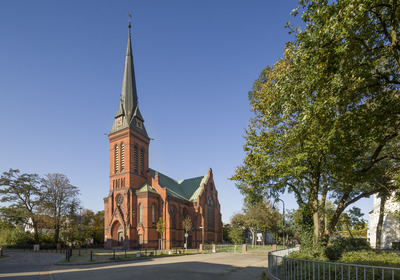 Eine rote Backsteinkirche mit einem hohen, spitzen Turm. Die Fassade ist verziert, und die Kirche steht umgeben von Bäumen. Der Himmel ist klar und blau, was eine ruhige Atmosphäre schafft.