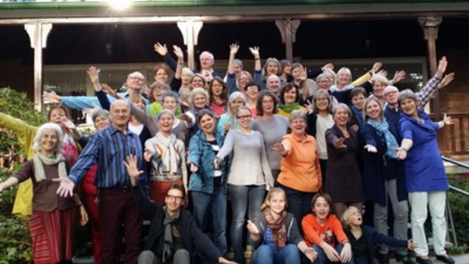 Eine große Gruppe von Menschen steht auf einer Treppe und lächelt in die Kamera. Viele halten die Arme nach oben oder zur Seite. Die Teilnehmer wirken fröhlich und vereint, und es ist eine freundliche, gesellige Atmosphäre spürbar.