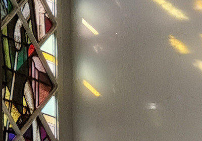 Buntglasfenster mit figürlichen Darstellungen, darunter eine stehende Person in einem grünen Gewand. Das Licht fällt durch die bunten Glasscheiben und erzeugt farbige Reflexionen auf der Wand.