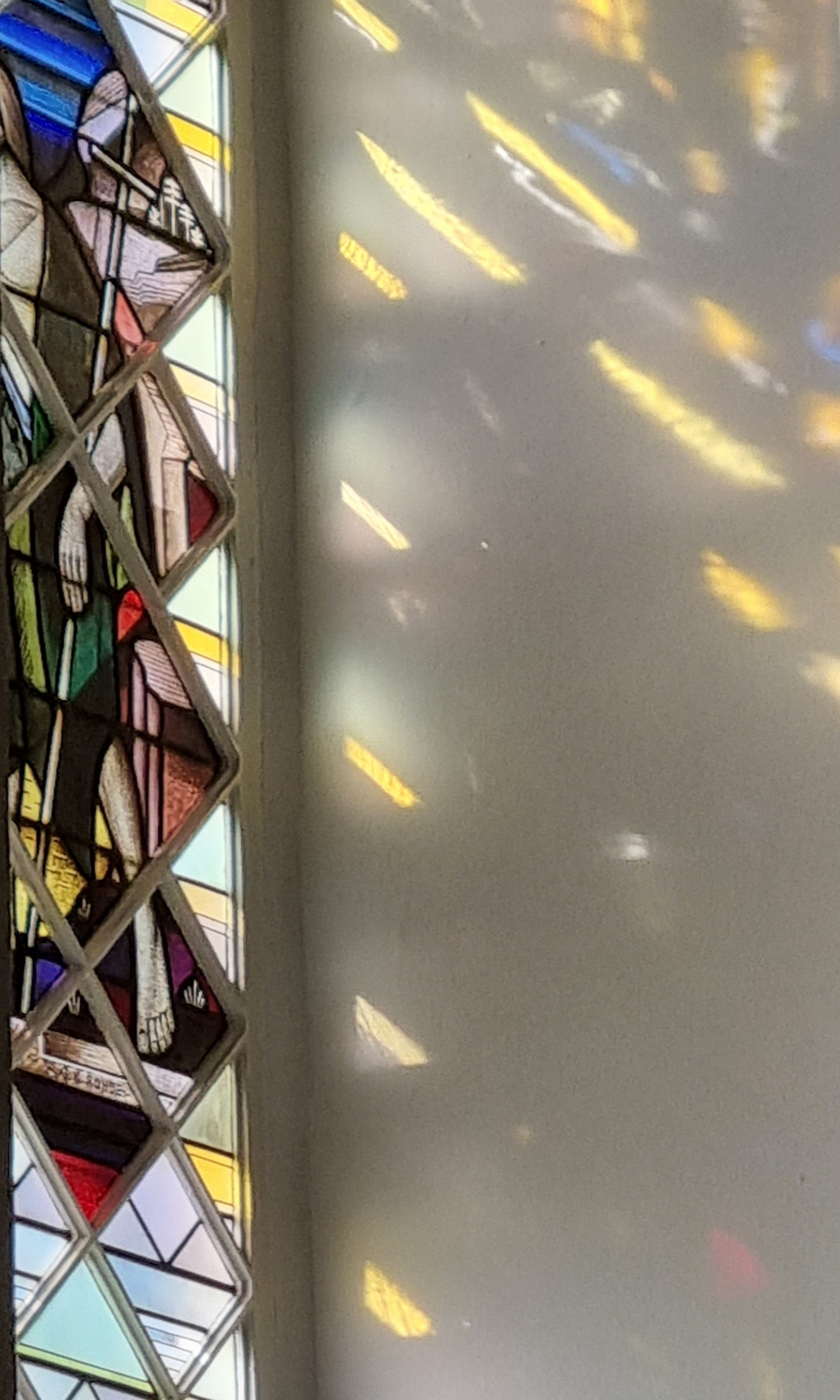 Buntglasfenster mit figürlichen Darstellungen, darunter eine stehende Person in einem grünen Gewand. Das Licht fällt durch die bunten Glasscheiben und erzeugt farbige Reflexionen auf der Wand.