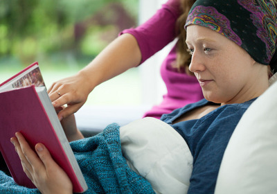 Das Symbolfoto zeigt eine lesende Frau im Bett eines Hospizes, mit einer Kopfbedeckung, weil sie offenbar keine Haare mehr hat
