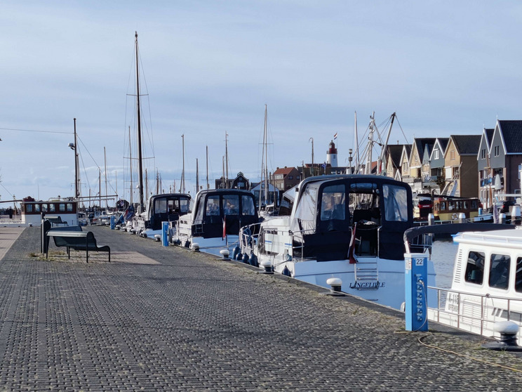 Ein Hafen mit mehreren Booten, die am Steg festgemacht sind. Die Liegestelle ist mit grauen Steinen gepflastert, und im Hintergrund sind verschiedene Gebäude und Mastanlagen sichtbar. Der Himmel ist klar und blau.