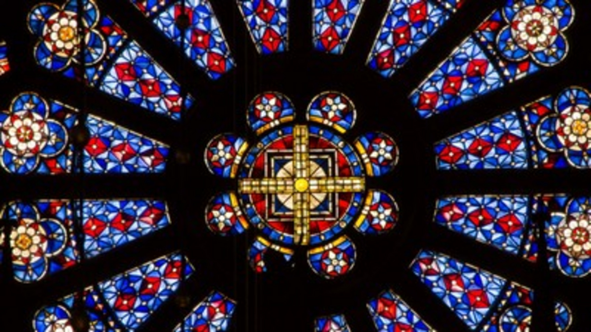 Ein buntes Glasfenster zeigt ein zentrales rosettenförmiges Design mit einem goldenen Kreuz in der Mitte. Um das Kreuz herum sind symmetrische Muster in Blau, Rot und Weiß angeordnet, die dem Fenster einen kunstvollen und spirituellen Charakter verleihen.