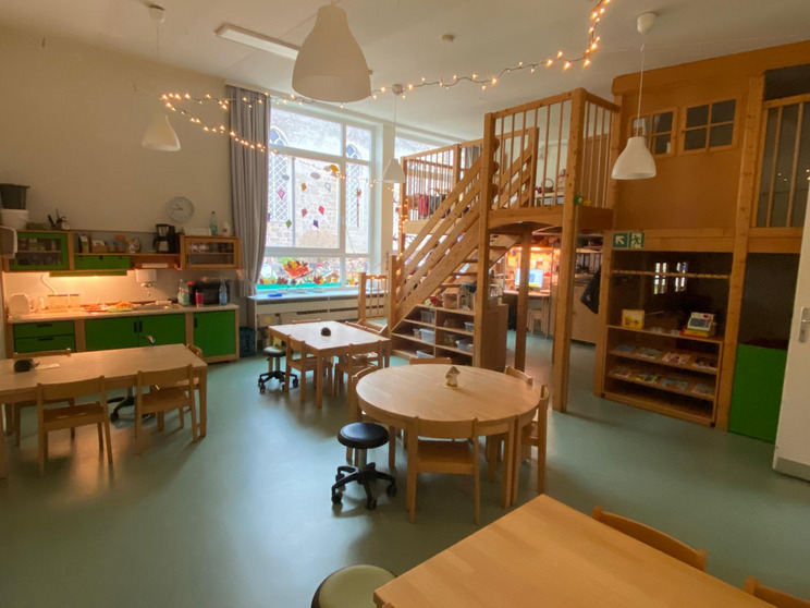 Ein heller Raum mit mehreren Holztischen und -stühlen. An der Wand befindet sich eine Küchenzeile mit einem Waschbecken. Eine Treppe führt zu einem Holzbereich im oberen Stockwerk. Große Fenster lassen Tageslicht herein, und die Wände sind mit bunten Dekorationen geschmückt.