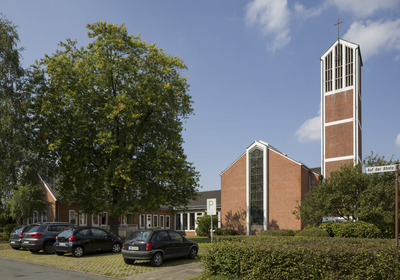 Das Bild zeigt eine moderne Kirche aus roten Ziegeln mit einem hohen Turm, der ein Kreuz trägt. Um die Kirche herum stehen mehrere Autos auf einem Parkplatz, und es sind einige Bäume und Sträucher zu sehen. Der Himmel ist blau mit vereinzelten Wolken.
