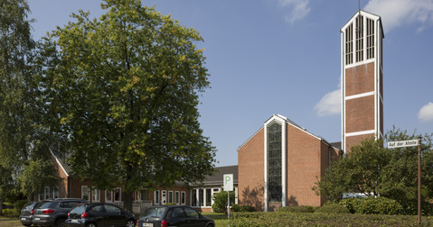 Das Bild zeigt eine moderne Kirche aus roten Ziegeln mit einem hohen Turm, der ein Kreuz trägt. Um die Kirche herum stehen mehrere Autos auf einem Parkplatz, und es sind einige Bäume und Sträucher zu sehen. Der Himmel ist blau mit vereinzelten Wolken.