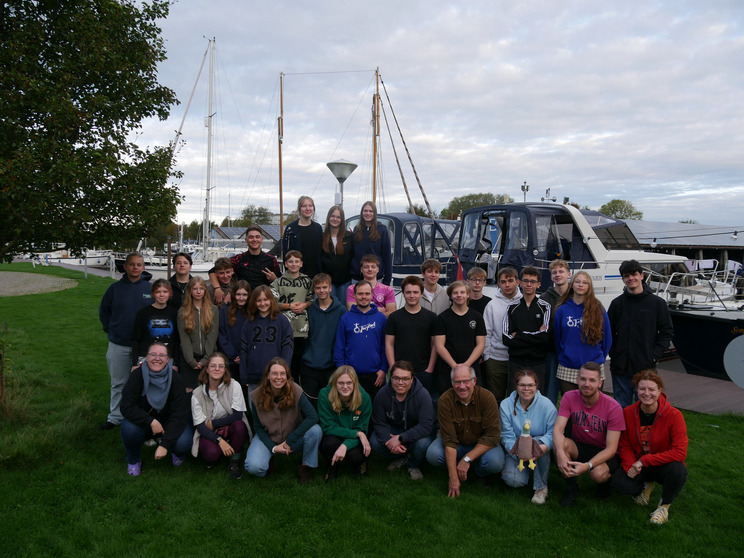 Gruppenfoto 2025 Eine Gruppe von etwa 30 Personen steht auf einer Wiese am Wasser. Im Hintergrund sind Boote und ein bewölkter Himmel zu sehen. Die Personen lächeln und posieren gemeinsam, einige sitzen, während andere stehen. Es handelt sich um eine freundliche und gesellige Atmosphäre.