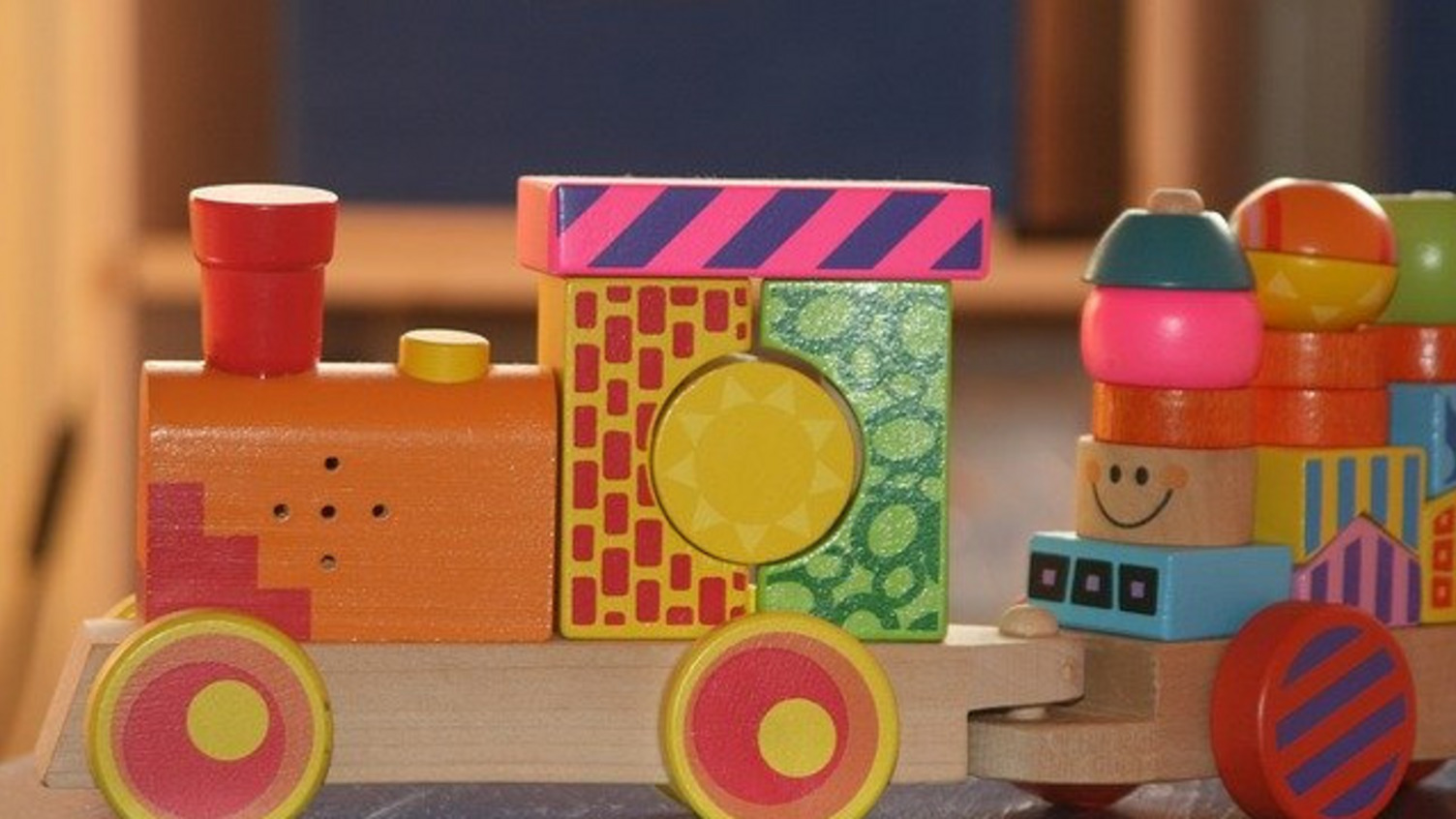 Ein buntes Spielzeug-Eisenbahn aus Holz mit verschiedenen geometrischen Formen, darunter Kreise und Quadrate. Die Lok hat einen roten Schornstein und leuchtende Farben, während der Waggon mit weiteren bunten Bausteinen dekoriert ist. Ideal für Kinder zum Spielen und Entdecken.