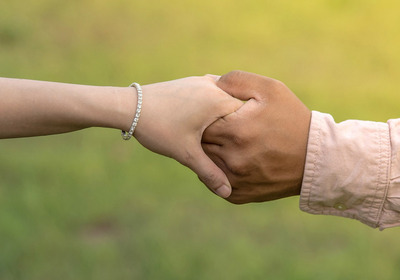 Zwei Hände greifen ineinander. Eine Hand trägt ein Armband, die andere steckt in einem weißen Hemd. Der Hintergrund ist grün und unscharf.
