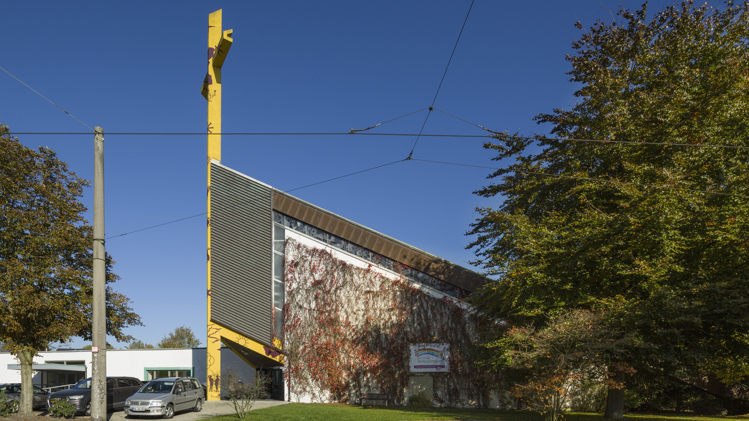Das Bild zeigt ein modernes Gebäude mit einer schräg verlaufenden Fassade, bedeckt mit Pflanzen. Im Vordergrund stehen Autos und Bäume. An der Fassade ist ein Schild angebracht. Der Himmel ist klar und blau.