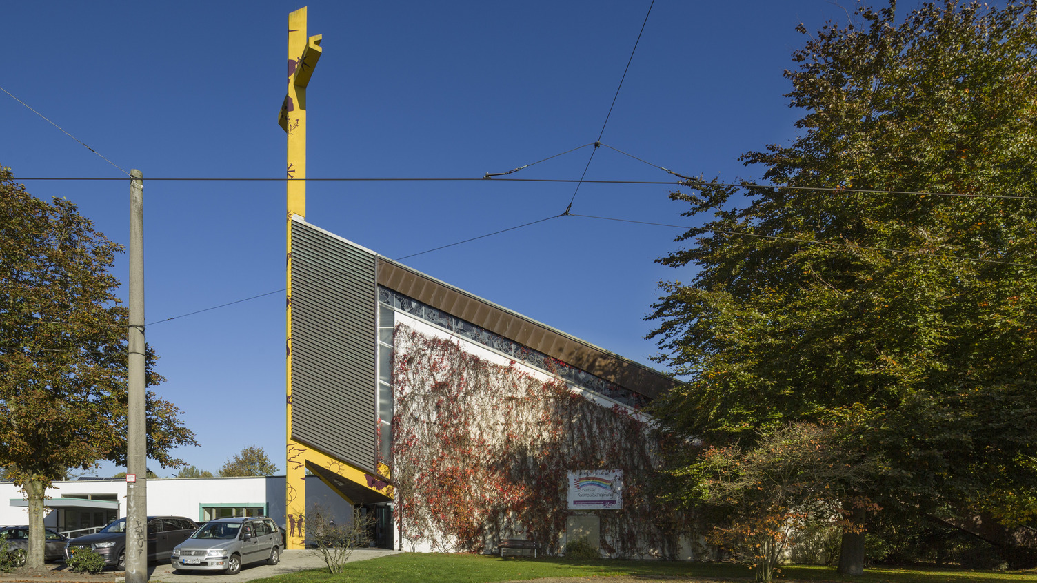 Das Bild zeigt ein modernes Gebäude mit einer schräg verlaufenden Fassade, bedeckt mit Pflanzen. Im Vordergrund stehen Autos und Bäume. An der Fassade ist ein Schild angebracht. Der Himmel ist klar und blau.