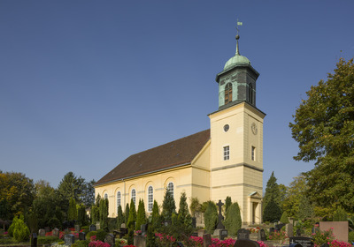 Eine gelbe Kirche mit einem markanten Turm steht umgeben von einem Friedhof mit Grabsteinen und bunten Blumen. Ein klarer blauer Himmel bildet den Hintergrund. Der Turm hat eine grüne Kuppel und Fenster.