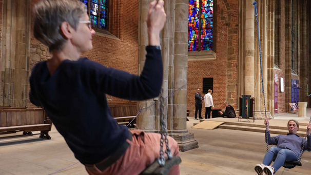 Das Foto zeigt eine Kunst-Installation in der Bremer Stadtkirche Unser Lieben Frauen. Zwei Frauen schaukeln miteinander im Takt.
