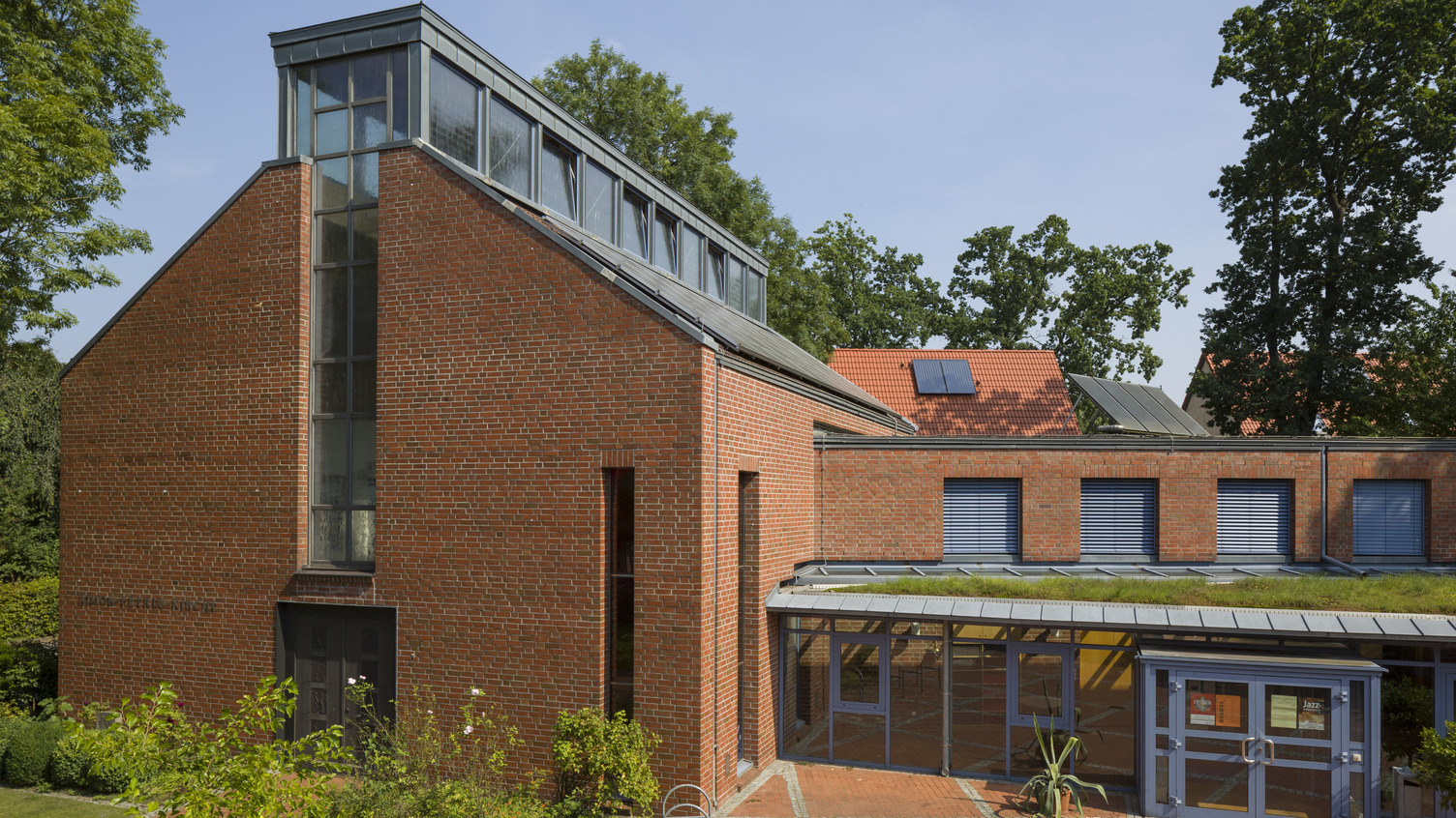 Ein modernes Gebäude aus roten Ziegeln mit einer großen Glasfront und einem flachen, begrünten Dach. Im Vordergrund ist ein schattiger Innenhof mit Pflastersteinen zu sehen. Bäume umgeben die Struktur, und der Himmel ist klar und blau.