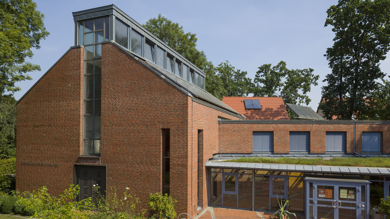 Ein modernes Gebäude aus roten Ziegeln mit einer großen Glasfront und einem flachen, begrünten Dach. Im Vordergrund ist ein schattiger Innenhof mit Pflastersteinen zu sehen. Bäume umgeben die Struktur, und der Himmel ist klar und blau.
