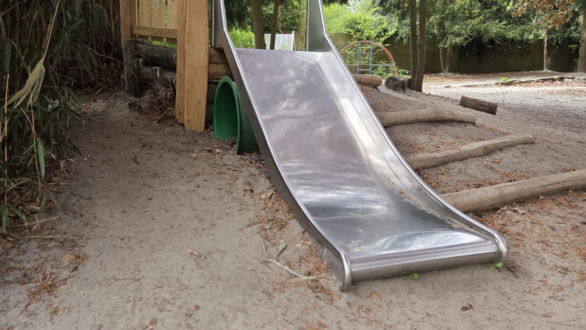 Eine Edelstahlrutsche steht an einem Spielplatz, umgeben von Bäumen und Sand. Im Hintergrund sind eine Holzstruktur und einige Baumstämme zu sehen. Die Atmosphäre ist ruhig, mit natürlichem Licht, das durch die Blätter fällt.