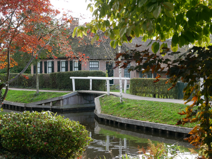 Giethoorn Brücke Ein ruhiger Kanal mit einem kleinen Holzsteg führt zu einem charmanten, reetgedeckten Haus. Umgeben von grüner Landschaft und buntem Herbstlaub schafft die Szenerie eine friedliche Atmosphäre.
