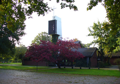 Eine Kirche mit einem hohen, modernen Glockenturm, auf dem ein Kreuz steht. Im Vordergrund blüht ein rotblättriger Baum, umgeben von einem gepflegten Garten. Weitere Bäume sind im Hintergrund sichtbar, während der Himmel klar und hell ist.