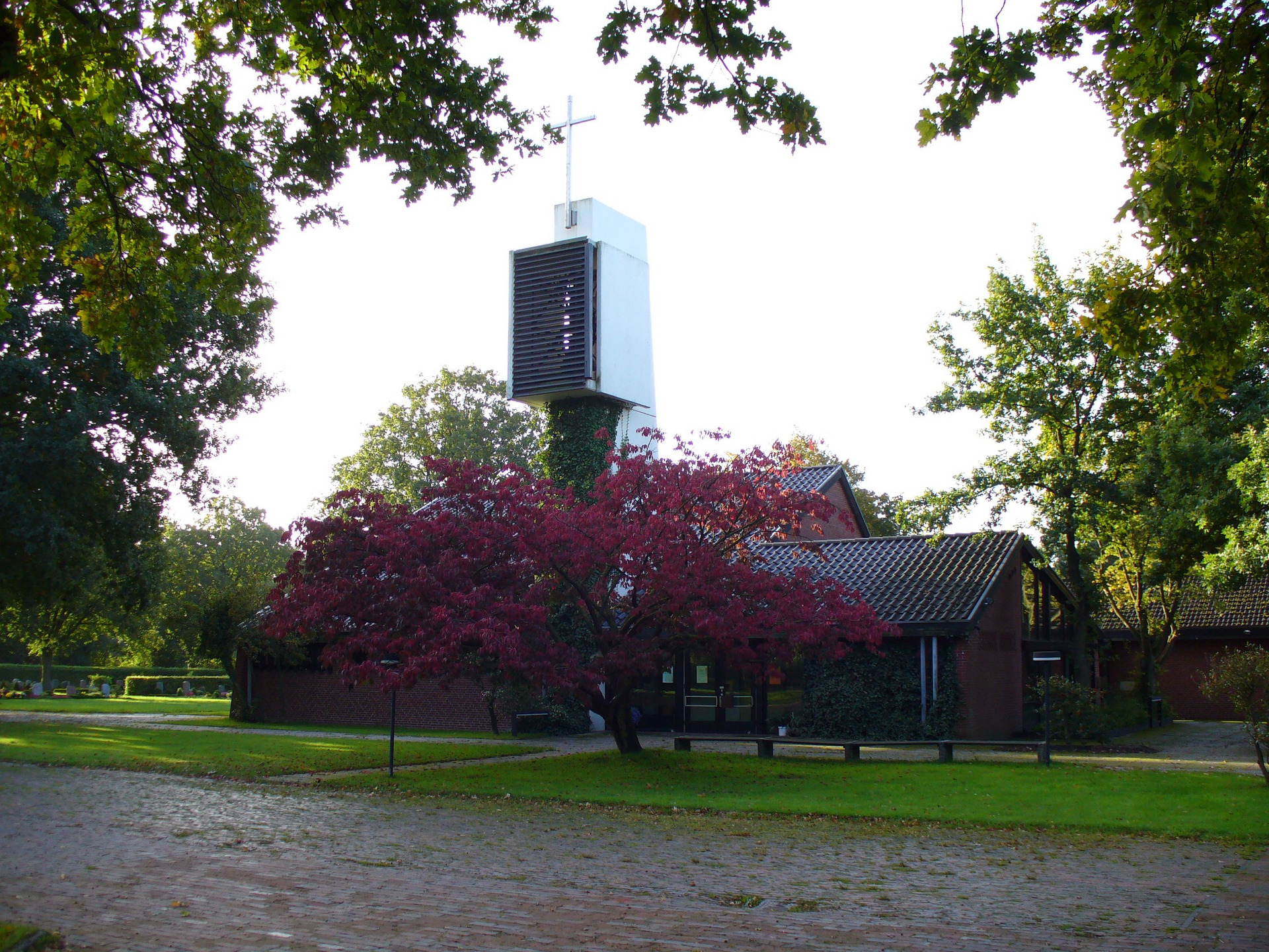 Eine Kirche mit einem hohen, modernen Glockenturm, auf dem ein Kreuz steht. Im Vordergrund blüht ein rotblättriger Baum, umgeben von einem gepflegten Garten. Weitere Bäume sind im Hintergrund sichtbar, während der Himmel klar und hell ist.