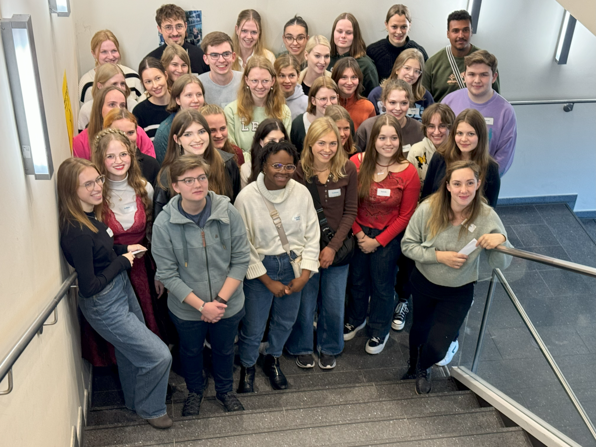 Gruppenfoto von etwa 40 Personen, die auf einer Treppe stehen. Die Gruppe besteht hauptsächlich aus jungen Erwachsenen, die freundlich lächeln und sich in lässiger Kleidung präsentieren. Einige tragen Namensschilder. Der Hintergrund zeigt eine helle Treppe mit einem grauen Boden.