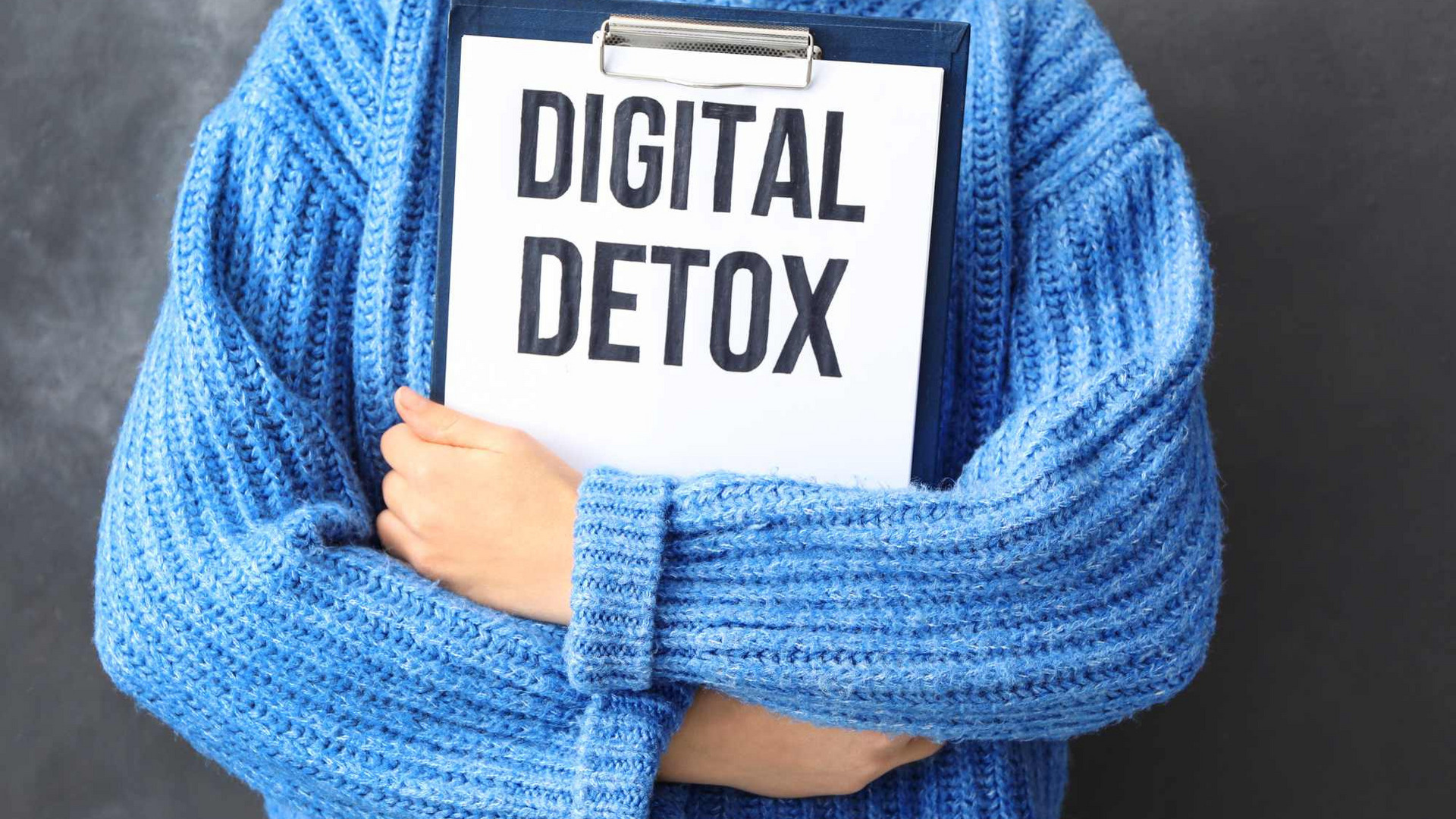 Eine Person trägt einen blauen Pullover und hält ein Schild mit der Aufschrift „DIGITAL DETOX“. Der Hintergrund ist graufarben. Der Fokus liegt auf der Botschaft, die digitale Auszeiten betont.