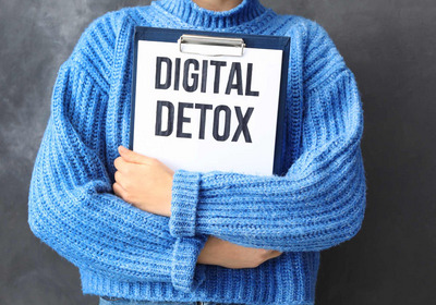 Eine Person trägt einen blauen Pullover und hält ein Schild mit der Aufschrift „DIGITAL DETOX“. Der Hintergrund ist graufarben. Der Fokus liegt auf der Botschaft, die digitale Auszeiten betont.