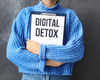 Eine Person trägt einen blauen Pullover und hält ein Schild mit der Aufschrift „DIGITAL DETOX“. Der Hintergrund ist graufarben. Der Fokus liegt auf der Botschaft, die digitale Auszeiten betont.