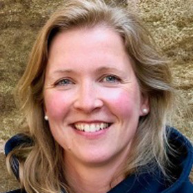 Porträt einer lächelnden Frau mit blonden Haaren, die in einem blauen Kapuzenpulli gekleidet ist. Der Hintergrund ist unscharf und zeigt eine natürliche, steinige Textur.