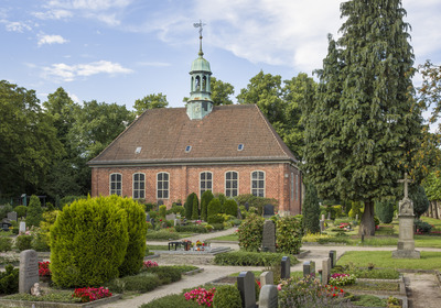 Das Bild zeigt eine rote Backsteinkirche mit einem kleinen grünen Turm, der aus der Mitte des Gebäudes ragt. Um die Kirche herum gibt es einen gepflegten Friedhof mit Grabsteinen und bunten Blumenbeeten. Im Hintergrund sind hohe Bäume und ein blauer Himmel zu sehen.
