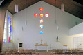 Der Innenraum einer Kirche mit einem einfachen Altar in der Mitte. Über dem Altar befindet sich ein Kreuz aus bunten Kirchenfenstern. Auf der linken Wand sind farbige Glasscheiben zu sehen. Der Raum hat eine ruhige, sakrale Atmosphäre. Holz- und Sitzbänke sind angeordnet.