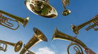 Sechs goldene Blasinstrumente, darunter Tuben und Trompeten, sind in einem Kreis angeordnet und blicken nach oben gegen einen strahlend blauen Himmel. Die Spiegelungen zeigen einen grünen Hintergrund und einige Personen.