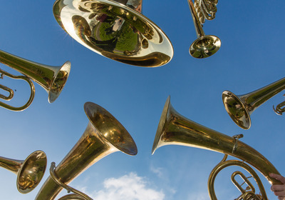 Sechs goldene Blasinstrumente, darunter Tuben und Trompeten, sind in einem Kreis angeordnet und blicken nach oben gegen einen strahlend blauen Himmel. Die Spiegelungen zeigen einen grünen Hintergrund und einige Personen.