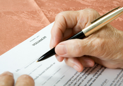 Eine Hand mit einem Schwarzstift schreibt auf einem Dokument mit der Überschrift „Vollmacht“. Die Schreibunterlage ist in einem warmen, einfarbigen Ton gehalten. Das Bild zeigt den Moment des Ausfüllens eines rechtlichen Dokuments.
