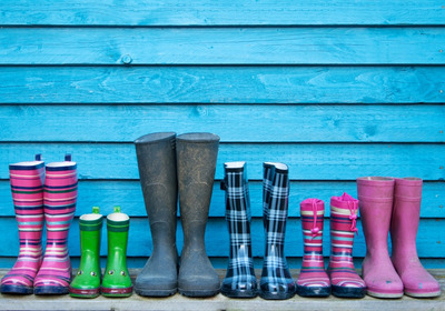 Mehrere Gummistiefel in verschiedenen Farben und Mustern stehen auf einem Holzbrett vor einer blauen Wand. Die Stiefel sind in Reihen angeordnet und variieren in Höhe und Design, einschließlich gestreiftem, kariertem und einfarbigem Muster.