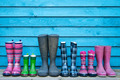 Mehrere Gummistiefel in verschiedenen Farben und Mustern stehen auf einem Holzbrett vor einer blauen Wand. Die Stiefel sind in Reihen angeordnet und variieren in Höhe und Design, einschließlich gestreiftem, kariertem und einfarbigem Muster.