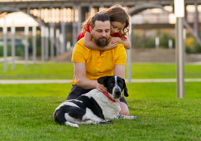 Ein Vater sitz auf einer Wiese mit seinem Kind und ihrem Hund.