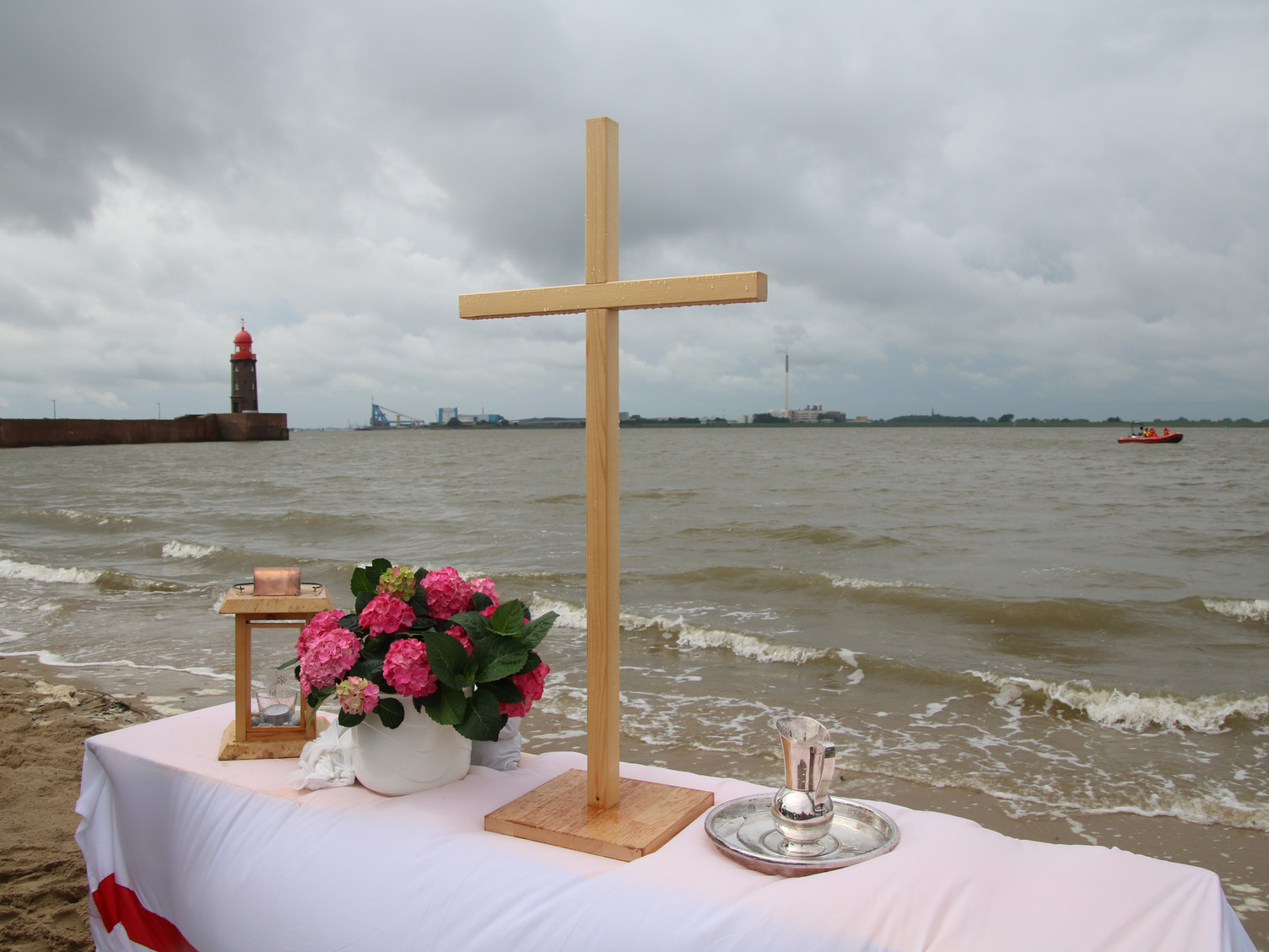 Ein Altar mit einem Holzkreuz steht an einem Strand. Im Hintergrund rauschen Meereswellen heran.