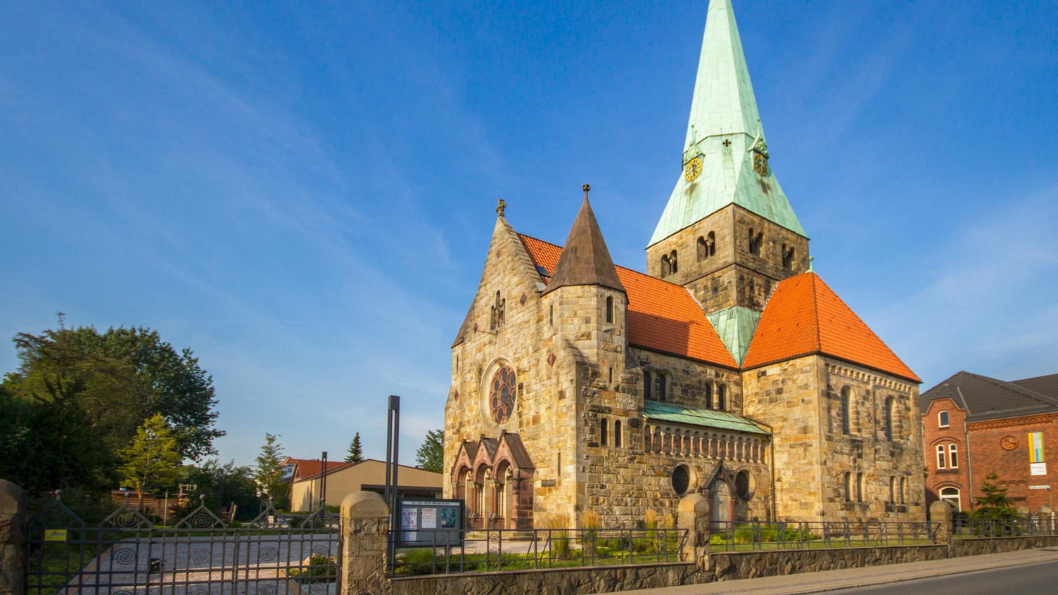 Eine historische Steinkirche mit einem hohen, grünen Turm und einem roten Satteldach. Die Fassade zeigt große Fenster und vielfältige architektonische Details. Im Vordergrund ist ein schmiedeeisernes Tor zu sehen, umgeben von Bäumen und modernen Gebäuden im Hintergrund.