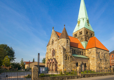 Eine historische Steinkirche mit einem hohen, grünen Turm und einem roten Satteldach. Die Fassade zeigt große Fenster und vielfältige architektonische Details. Im Vordergrund ist ein schmiedeeisernes Tor zu sehen, umgeben von Bäumen und modernen Gebäuden im Hintergrund.