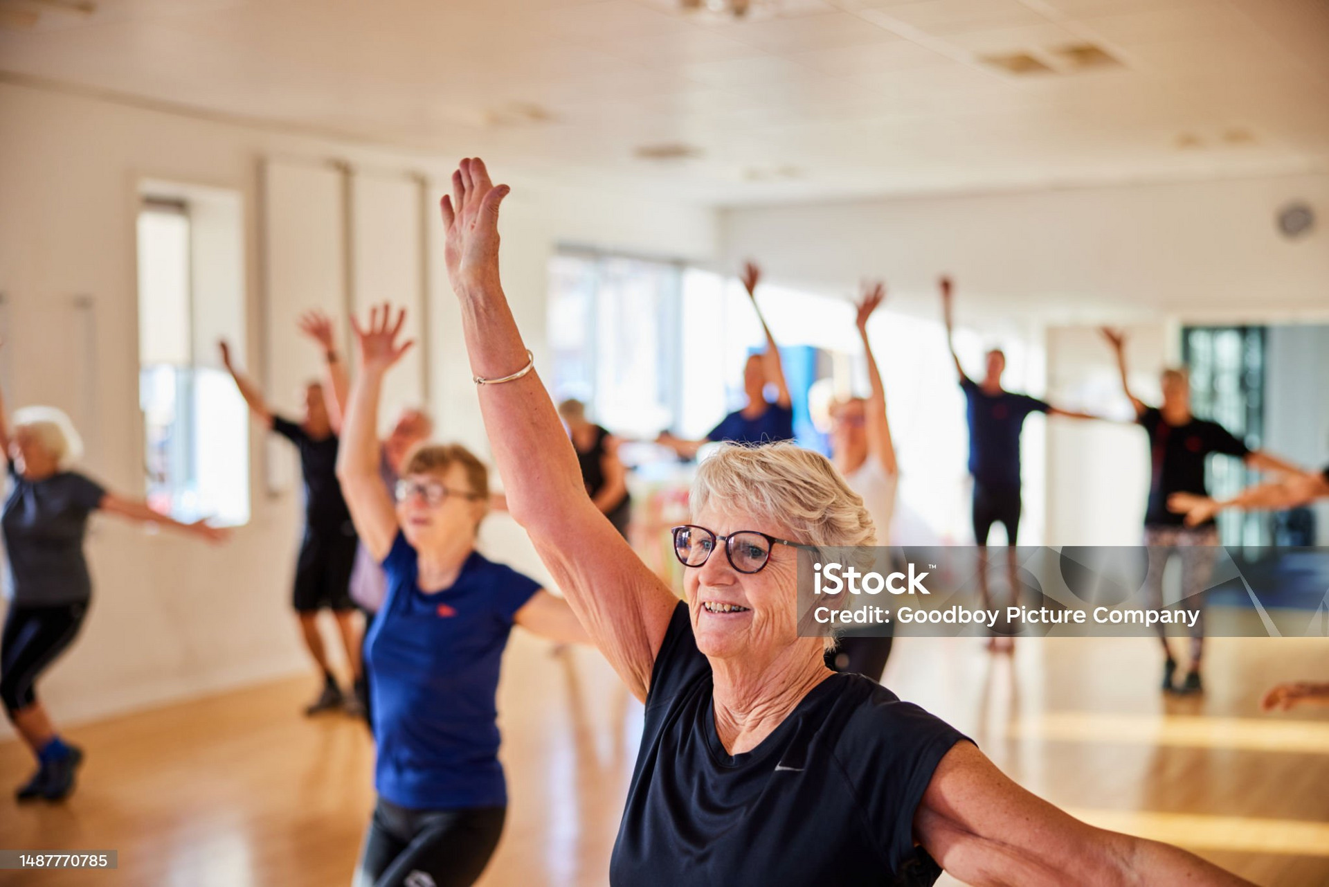 Eine Gruppe älterer Menschen steht in einem hellen Raum und macht gemeinsam Fitnessübungen. Sie lächeln und heben die Arme, während sie sich rhythmisch bewegen. Die Atmosphäre wirkt positiv und energiegeladen.