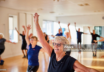 Eine Gruppe älterer Menschen steht in einem hellen Raum und macht gemeinsam Fitnessübungen. Sie lächeln und heben die Arme, während sie sich rhythmisch bewegen. Die Atmosphäre wirkt positiv und energiegeladen.
