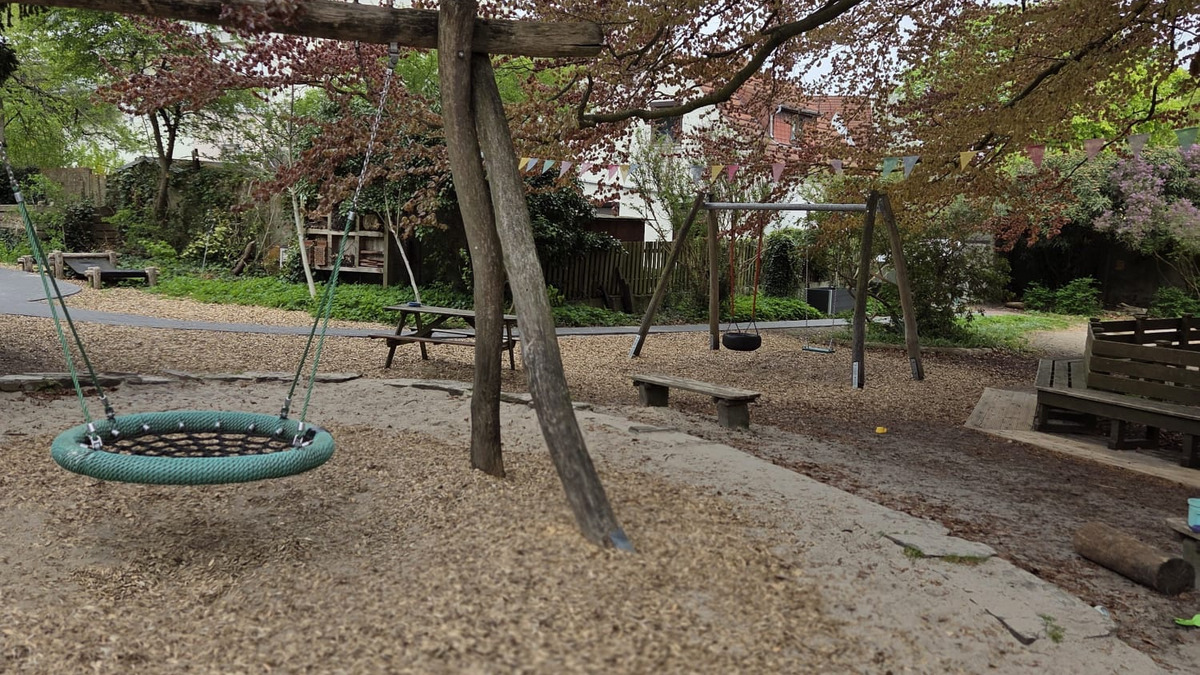 Ein Spielplatz mit einer großen, runden Schaukel und einer klassischen Schaukel aus Holz. Im Hintergrund sind Bänke und ein Spielbereich sichtbar. Über dem Spielplatz befinden sich Blütenzweige eines Baumes. Der Boden ist mit Holzspänen bedeckt.