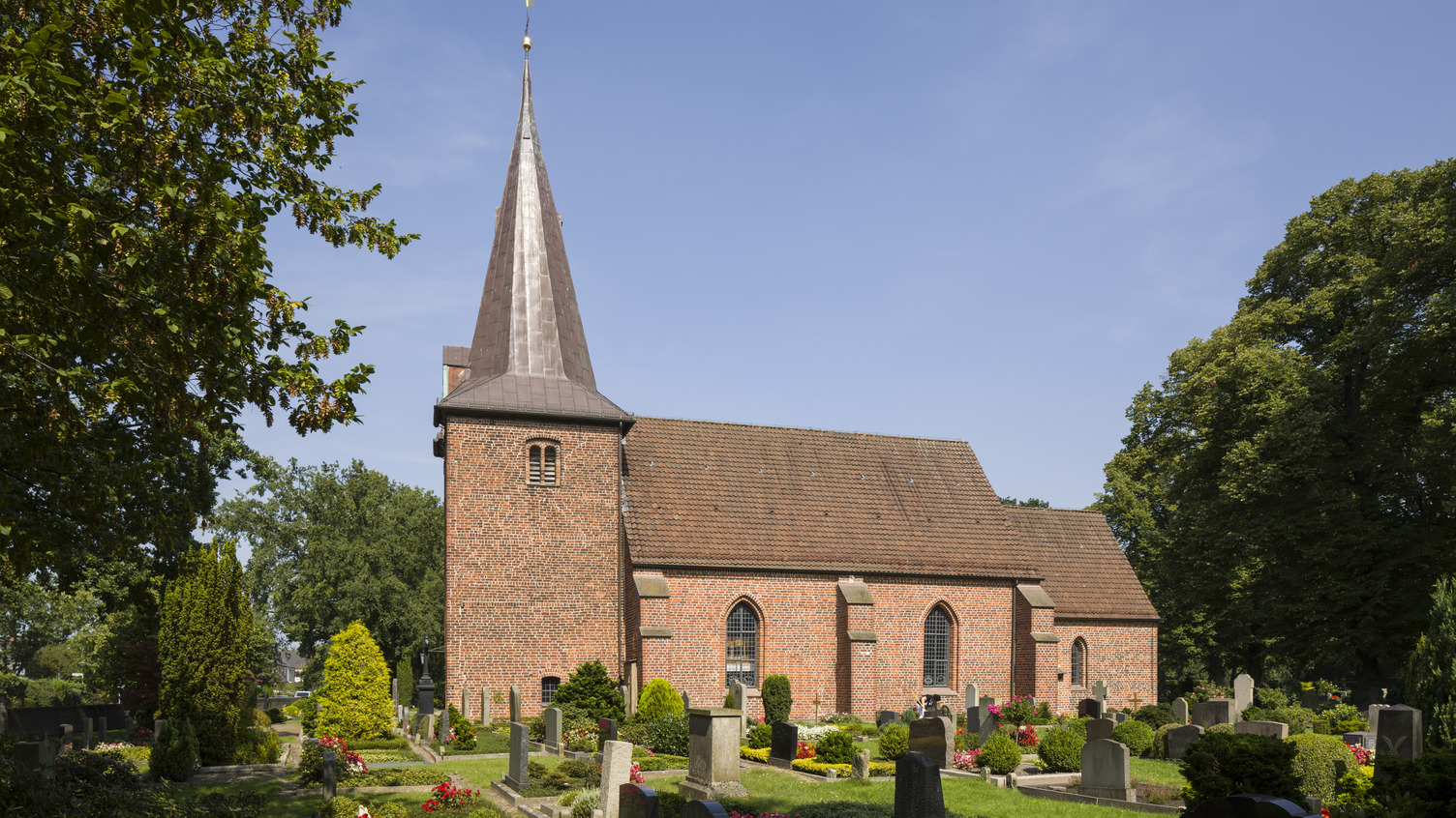 Eine rote Backsteinkirche mit einem spitzen Turm steht inmitten eines Friedhofs. Rund um die Kirche sind Gräber mit Steinen und bunten Blumenbeeten angeordnet. Im Hintergrund sind Bäume sichtbar und der Himmel ist klar.