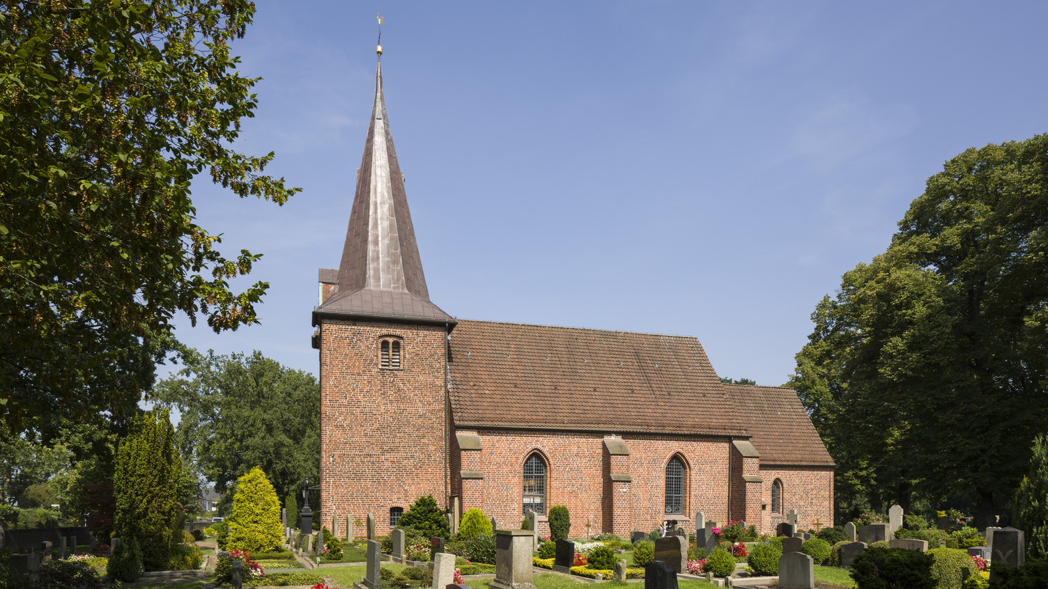Eine rote Backsteinkirche mit einem spitzen Turm steht inmitten eines Friedhofs. Rund um die Kirche sind Gräber mit Steinen und bunten Blumenbeeten angeordnet. Im Hintergrund sind Bäume sichtbar und der Himmel ist klar.
